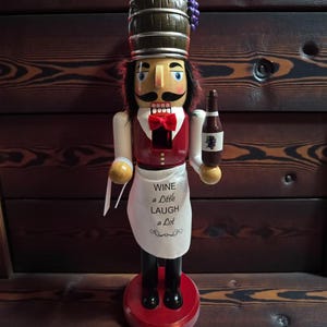 Peut inclure: Casse-noisette en bois en forme de serveur de vin, portant un gilet rouge, un tablier blanc et un chapeau en forme de tonneau. Le tablier indique "Wine a Little Laugh a Lot". Le casse-noisette tient une bouteille de vin.