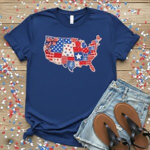 May include: A navy blue t-shirt featuring a patchwork design of the United States map in red, white, and blue patterns. The shirt is paired with denim shorts and brown sandals, set against a wooden background scattered with confetti.