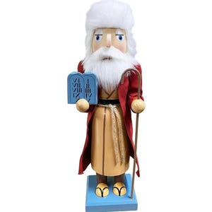 May include: A wooden nutcracker in the form of a biblical figure, wearing a white fur hat, a red robe, and a tan tunic. The figure holds blue tablets with Roman numerals and a wooden staff. The nutcracker stands on a blue base.