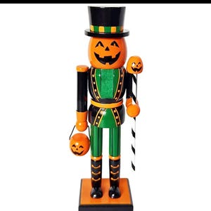 May include: A wooden nutcracker figure featuring a carved orange pumpkin head with a black jack-o'-lantern face, wearing a black top hat with a green band. The figure wears a green and black uniform, holds a small pumpkin bucket, and a staff.