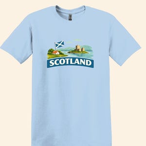 May include: Light blue t-shirt featuring a graphic of a Scottish landscape with a castle, flag, and the word "SCOTLAND" in white text on a blue banner. The design includes a small house and mountains.