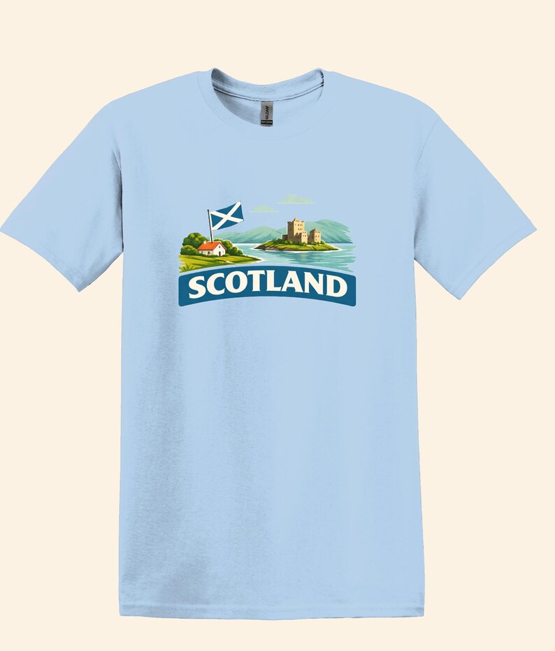 May include: Light blue t-shirt featuring a graphic of a Scottish landscape with a castle, flag, and the word "SCOTLAND" in white text on a blue banner. The design includes a small house and mountains.