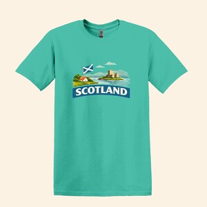 May include: Teal t-shirt featuring a graphic of a Scottish landscape with a castle, flag, and the word "SCOTLAND" in white on a blue banner. The design includes a small house and rolling hills.