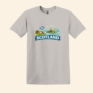 May include: A light gray t-shirt featuring a graphic of Scotland. The graphic includes a Scottish flag, a small house, a castle on an island, and the word "SCOTLAND" in a blue banner.