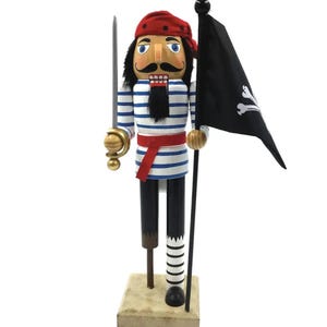 May include: A wooden nutcracker dressed as a pirate, wearing a red bandana, a blue and white striped shirt, and a red belt. It has a black beard, holds a silver sword in one hand, and carries a black flag with a white skull and crossbones design.