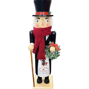 Könnte beinhalten: Eine hölzerne Nussknacker-Figur mit schwarzem Zylinder mit rotem Band, dunkelrotem Strickschal und schwarzem Mantel. Die Figur hält einen Holzstock und einen kleinen grünen Kranz mit roter Schleife. Eine weiße Schürze trägt den Text I WILL HONOR CHRISTMAS.