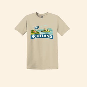 May include: A tan t-shirt featuring a graphic of a Scottish landscape with a castle, a flag, and the word "SCOTLAND" in a blue banner. The design includes a small house and rolling hills.