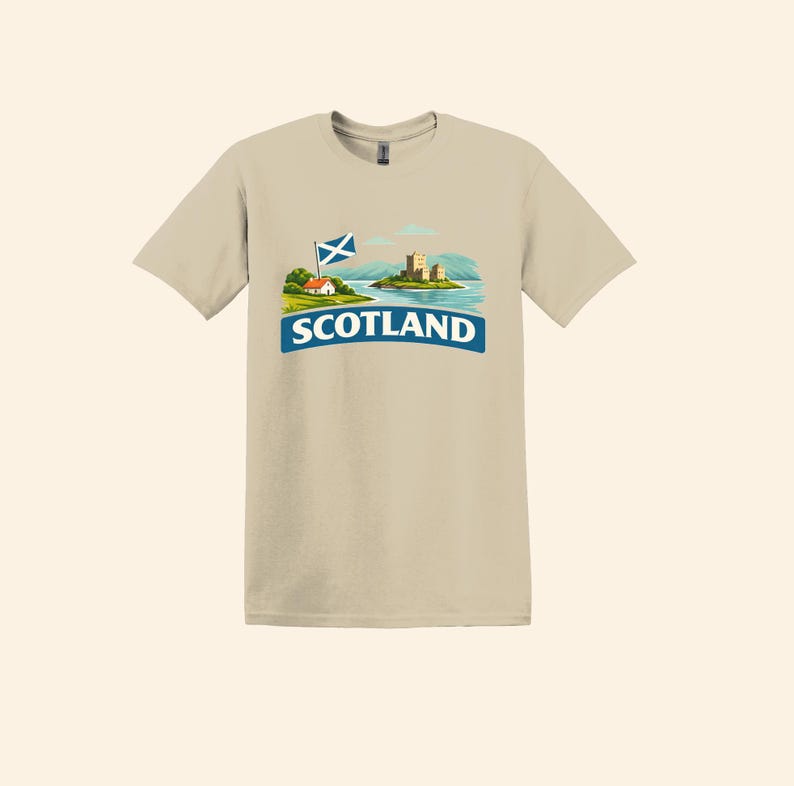 May include: A tan t-shirt featuring a graphic of a Scottish landscape with a castle, a flag, and the word "SCOTLAND" in a blue banner. The design includes a small house and rolling hills.
