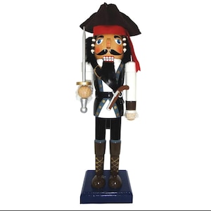 May include: A wooden pirate nutcracker figurine. The pirate wears a black hat, red bandana, and a black vest over a white shirt. He holds a sword and a wooden ball. The base is dark blue.