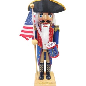 Puede incluir: Un cascanueces de madera con forma de soldado, sosteniendo una bandera estadounidense y un tambor. El cascanueces lleva un abrigo azul con detalles rojos, un sombrero negro y botas negras. El tambor tiene el texto "America 250". La base dice "Est. 1776".