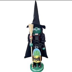 May include: A wooden nutcracker figure styled as a witch with green skin, wearing a tall black pointed hat and a black cape. The figure holds a broomstick and features a painted graphic on its torso depicting a yellow road leading to a green city.
