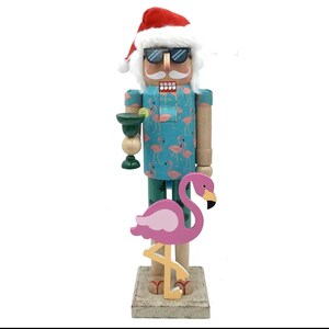 Könnte beinhalten: Ein Holz-Nussknacker-Figur mit Weihnachtsmütze und Sonnenbrille, die eine Margarita hält. Der Nussknacker hat ein blaues Hemd mit rosa Flamingomustern und grüne Hosen. Ein rosa Flamingo steht vor dem Nussknacker.