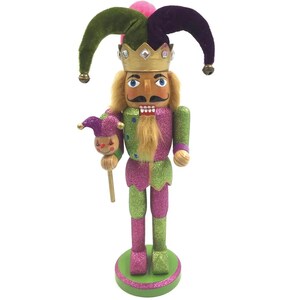 May include: A jester nutcracker figurine featuring a gold crown with faux gems, a long tan beard, and a glittery purple and lime green outfit. The figure wears a two-toned velvet hat and holds a small jester-faced scepter in one hand.
