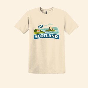 May include: Cream-colored t-shirt featuring a graphic of a Scottish landscape. The design includes a castle on an island, a small house, and the text "SCOTLAND" in a blue banner, with the Scottish flag.