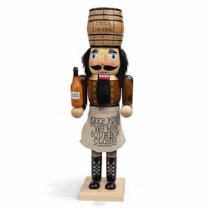 May include: Wooden nutcracker figurine with a barrel hat and a bottle of bourbon. The nutcracker has a black mustache and beard, and wears an apron that says "Keep Your Friends Close and Your Bourbon Closer."
