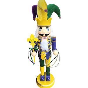 May include: A decorative nutcracker in a jester costume with a yellow, purple, and green hat. The nutcracker has a yellow crown, a white beard, and holds a yellow fleur-de-lis staff. It is adorned with beads and stands on a yellow base.