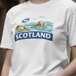 May include: White t-shirt featuring a scenic illustration of Scotland. The design includes a blue and white Scottish flag, a castle, and the word "SCOTLAND" in a blue banner. The shirt is made of a soft, comfortable material.