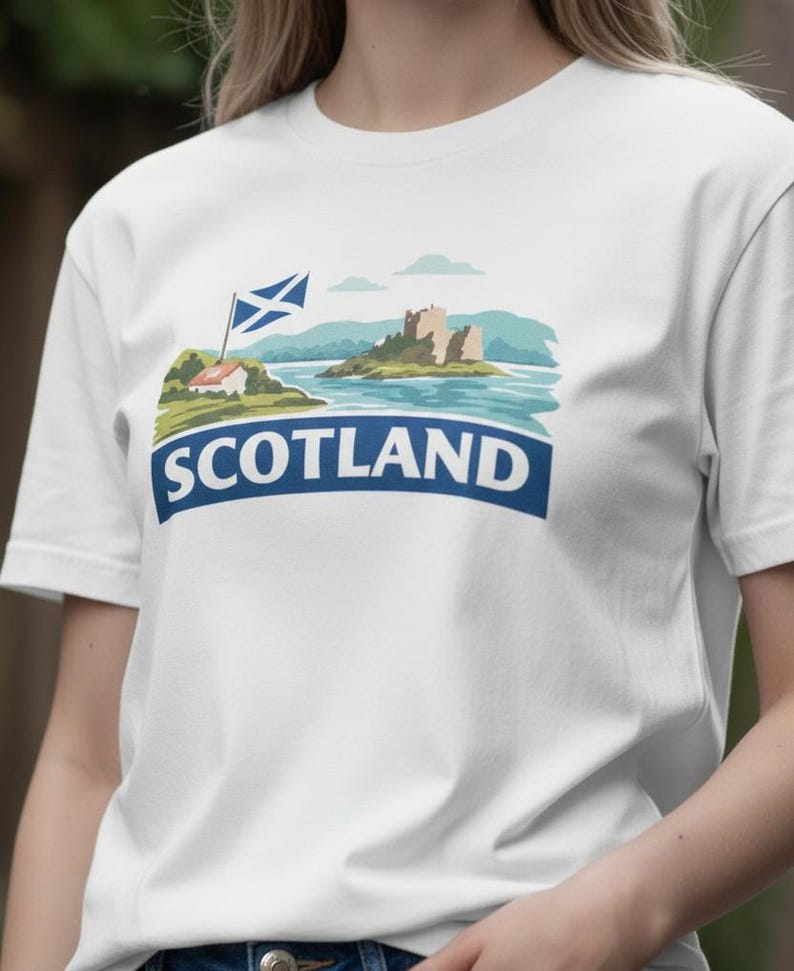 May include: White t-shirt featuring a scenic illustration of Scotland. The design includes a blue and white Scottish flag, a castle, and the word "SCOTLAND" in a blue banner. The shirt is made of a soft, comfortable material.