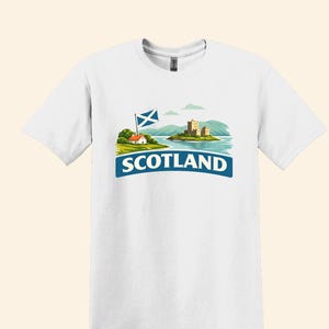 May include: White t-shirt featuring a graphic of Scotland. The design includes a Scottish flag, a small house, a castle on an island, and the word "SCOTLAND" in a blue banner. The shirt is a short-sleeved crew neck.