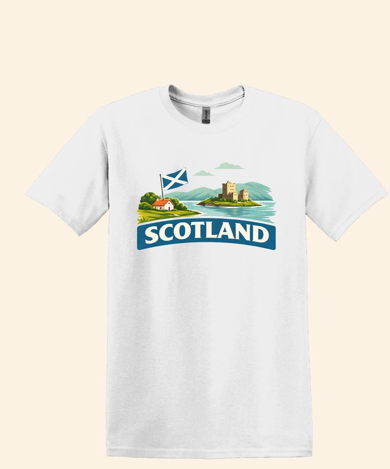 May include: White t-shirt featuring a graphic of Scotland. The design includes a Scottish flag, a small house, a castle on an island, and the word "SCOTLAND" in a blue banner. The shirt is a short-sleeved crew neck.