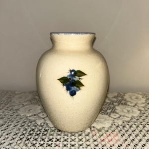 May include: A cream-colored ceramic vase with a slightly flared rim and a rounded body. The vase features a hand-painted design of blue blueberries and green leaves. The rim has a blue trim.