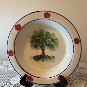 Puede incluir: Un cuenco de cerámica blanco con un borde azul y un diseño de árbol pintado. El árbol tiene hojas verdes y frutos rojos. Frutos rojos acentúan el borde. El cuenco está sobre un soporte negro.