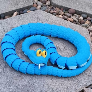 May include: A coiled blue toy snake with yellow eyes and a white fang. The snake is made of segmented pieces, giving it a textured appearance. It rests on a gray stone surface.