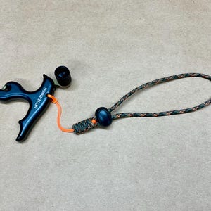 May include: A dark blue archery release aid with the text "SPOT-HOGG" printed on the side. It has an orange cord and a camouflage-patterned rope loop. The release aid is designed for use with a bow.
