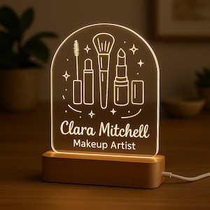 Könnte beinhalten: Ein personalisiertes LED-Nachtlicht für Maskenbildner. Die transparente Acrylplatte zeigt geätzte Make-up-Tools und den Text "Clara Mitchell Makeup Artist". Es steht auf einem Holzsockel und strahlt ein warmes Licht aus.