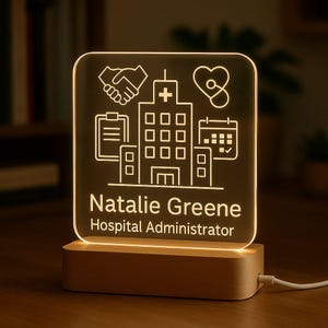Może przedstawiać: Spersonalizowana lampka nocna LED z drewnianą podstawą. Przezroczysty panel akrylowy przedstawia motyw szpitalny z ikonami i imieniem "Natalie Greene, Hospital Administrator" w kolorze białym. Światło emituje ciepłą poświatę.