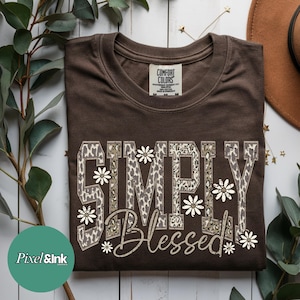 May include: A folded brown t-shirt with the words "SIMPLY BLESSED" in a decorative font. The letters are filled with a leopard print pattern and adorned with white daisy flowers. The shirt has a Comfort Colors tag.