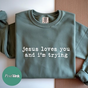 May include: Sage green sweatshirt with the text "jesus loves you and I'm trying" in white. The sweatshirt has a crew neck and long sleeves. The Pixel & Ink logo is in the bottom left corner.
