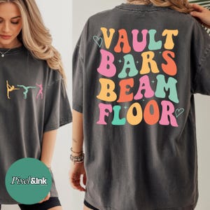 Puede incluir: Camiseta gris oscuro con texto y gráficos coloridos. La parte posterior de la camiseta muestra las palabras "VAULT BARS BEAM FLOOR" en una fuente retro. La parte delantera presenta figuras de gimnastas en naranja, turquesa y rosa.