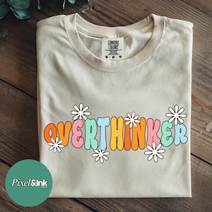 May include: A beige Comfort Colors t-shirt with the word "OVERTHINKER" in colorful, bubble letters, accented with white daisy graphics. The shirt is folded on a wooden surface.