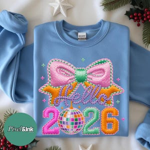 May include: A light blue sweatshirt with a colorful embroidered design. The design includes a pink bow, the words "Hello 2026", and a disco ball. The numbers "2026" are in pink, green, and orange. The sweatshirt is made of cotton.
