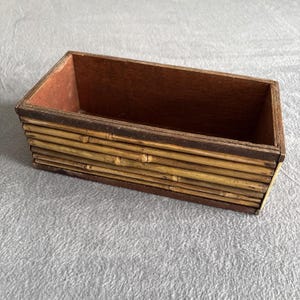 May include: Rectangular wooden box with a bamboo exterior. The box has a dark brown interior and a frame around the top. The bamboo is a light yellow color.