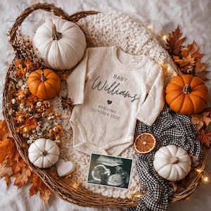 Op de afbeelding: Een herfstthema baby aankondiging. Een crèmekleurige baby-onesie met de tekst "Baby Williams Arriving October 2026" is omringd door pompoenen, bladeren en een echografiebeeld, alles gerangschikt in een gevlochten mand.