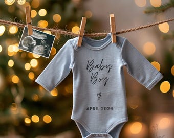 May include: A light blue baby onesie with the words "Baby Boy" and "April 2026" printed on it, hanging on a clothesline with a sonogram image. The background features blurred bokeh lights.