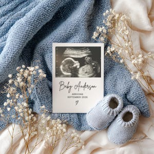 May include: A baby announcement card with an ultrasound image, the text "Baby Anderson ARRIVING SEPTEMBER 2026", and a heart symbol. Light blue knitted baby booties are next to the card, all on a blue knitted blanket with white flowers.