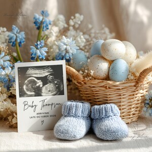 May include: A baby announcement featuring a sonogram image, text reading "Baby Thompson ARRIVING LATE OCTOBER 2026," and a heart symbol. Light blue knitted baby booties sit in front of the announcement. A wicker basket holds speckled eggs, and blue and white flowers are in the background.