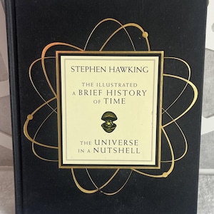 May include: A black hardcover book titled The Illustrated A Brief History of Time and The Universe in a Nutshell by Stephen Hawking. The cover features a gold atomic orbital design surrounding a central cream-colored rectangular label.