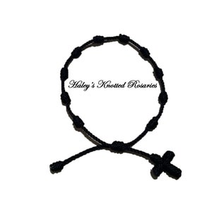 May include: A black knotted rosary bracelet with a small black cross charm. The bracelet is made of black cord and has a simple design. "Haley's Knotted Rosaries" is written in white text on a white background.