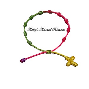 May include: A knotted rosary bracelet with green, pink, and yellow cord. The bracelet has a small yellow cross charm.