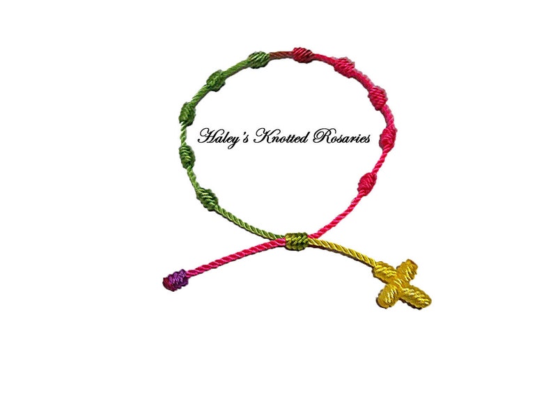 May include: A knotted rosary bracelet with green, pink, and yellow cord. The bracelet has a small yellow cross charm.
