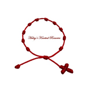 Peut inclure: Bracelet de prière en corde rouge nouée avec un pendentif en forme de croix. "Haley's Knotted Rosaries" est écrit sur le bracelet.
