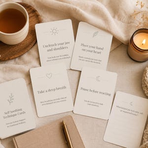 May include: A cup of tea, a lit candle, and several cream-colored cards with self-care prompts are arranged on a beige cloth. The cards have simple line drawings and phrases like "Take a deep breath" and "Pause before reacting."