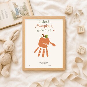 Può includere: Una stampa incorniciata con il testo Cutest Pumpkin in the Patch sopra un'impronta di mano arancione a forma di zucca con un gambo verde. Sotto l'impronta, il testo recita Made with little hands & lots of love, con linee per By e Date.