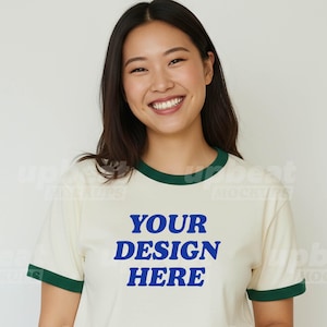 Puede incluir: Camiseta ringer color crema con ribetes verdes y el texto azul "YOUR DESIGN HERE". Esta camiseta es una maqueta, ideal para mostrar diseños personalizados. La modelo lleva vaqueros oscuros.