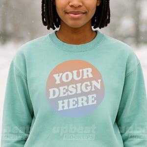Puede incluir: Una sudadera de color verde azulado claro con cuello redondo y mangas largas. La parte delantera presenta un diseño circular con el texto "YOUR DESIGN HERE" en blanco. El diseño está sobre un degradado de naranja, rosa y azul.