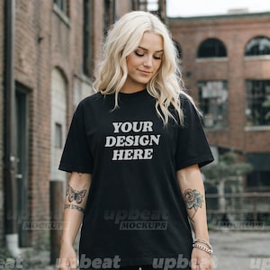 Puede incluir: Camiseta negra con el texto "YOUR DESIGN HERE" en blanco. La camiseta es una maqueta, perfecta para mostrar diseños personalizados. La modelo lleva pantalones cortos negros. El fondo es un edificio de ladrillo.